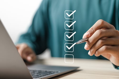 Close-up of a person holding a pen near a laptop, with a vertical series of digital checkboxes overlaid on the image, most marked with checkmarks to indicate completed tasks.