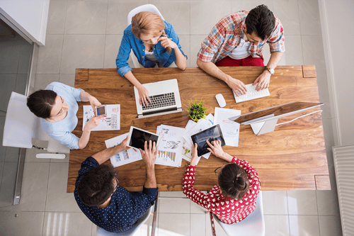 Overhead view of a team working around a wooden table, using laptops, tablets, and paper documents with charts and graphs, alongside a smartphone and desktop monitor.