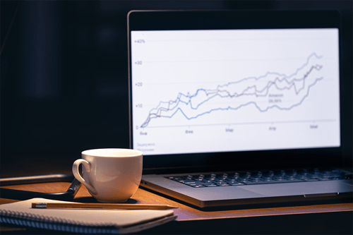 Laptop on a desk displaying a line chart with multiple upward-trending data lines, alongside a coffee cup, notebook, and pencil in a dimly lit workspace.