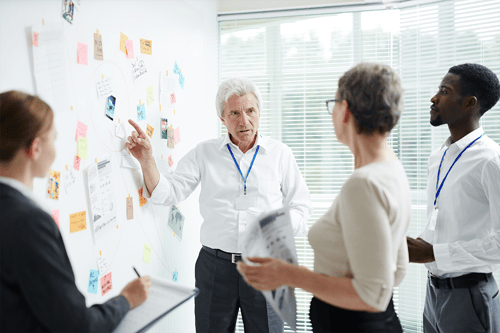 Group of professionals gathered in an office meeting, with one person pointing at a whiteboard covered in sticky notes, diagrams, and images while others review documents and take notes.